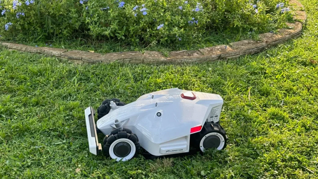  robotic lawn mower, labeled as a Mammotion LUBA AWD 5000, sits on a lush green lawn. In the background is a brick fence.