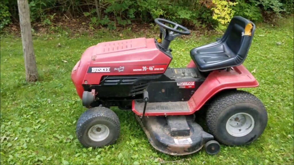 Red Huskee Yard Man tractor mower with black seat, parked on a green field next to a tree.