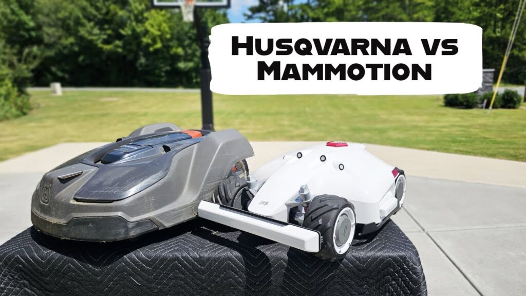 Two robotic lawn mowers, a Mammotion Luba AWD 5000 and a Husqvarna Automower, sit on a metal table in front of a basketball court. Text on the image reads "HUSQVARNA VS MAMMOTION".