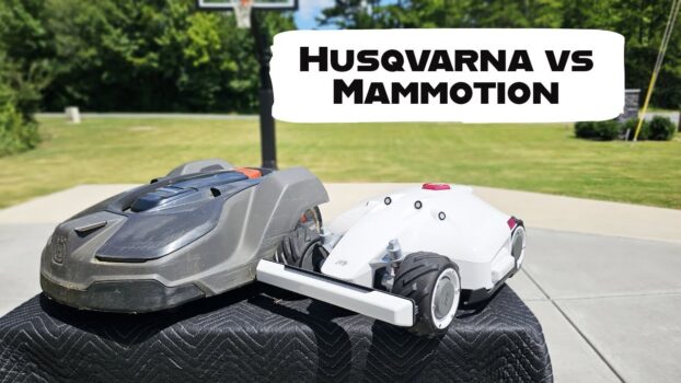 Two robotic lawn mowers sit side-by-side on a metal table. The text "HUSQVARNA VS MAMMOTION" is overlaid on the image. A Mammotion Luba AWD 5000 is on the left and a Husqvarna Automower is on the right.