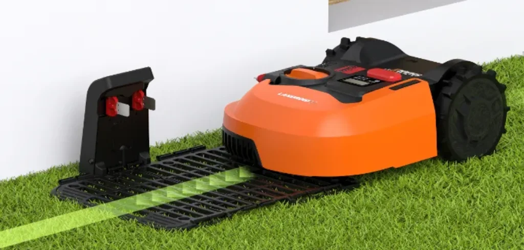 An orange and black robotic lawn mower with a rounded rectangular base and two protruding wheels on either side. It has a small black antenna on top and a grass trimmer underneath. The mower is cutting a line in a lush green lawn.